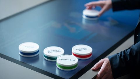 On a table with a scan and touch function, there are five cylinder-shaped control objects with crime prevention keywords on them. A person moves one of them with their hand.